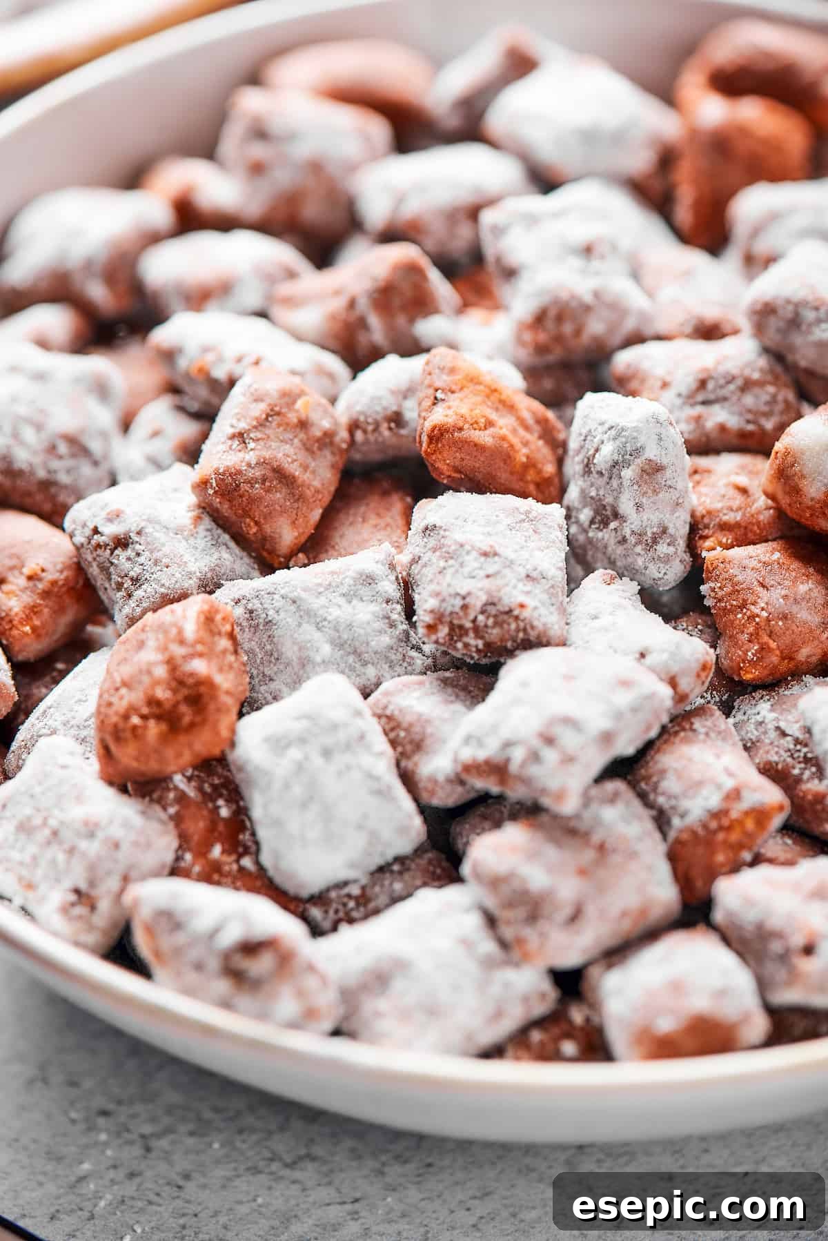 Close-up shot of puppy chow in a white bowl, to show the texture. Some pieces are fully coated in powdered sugar, while others are not.