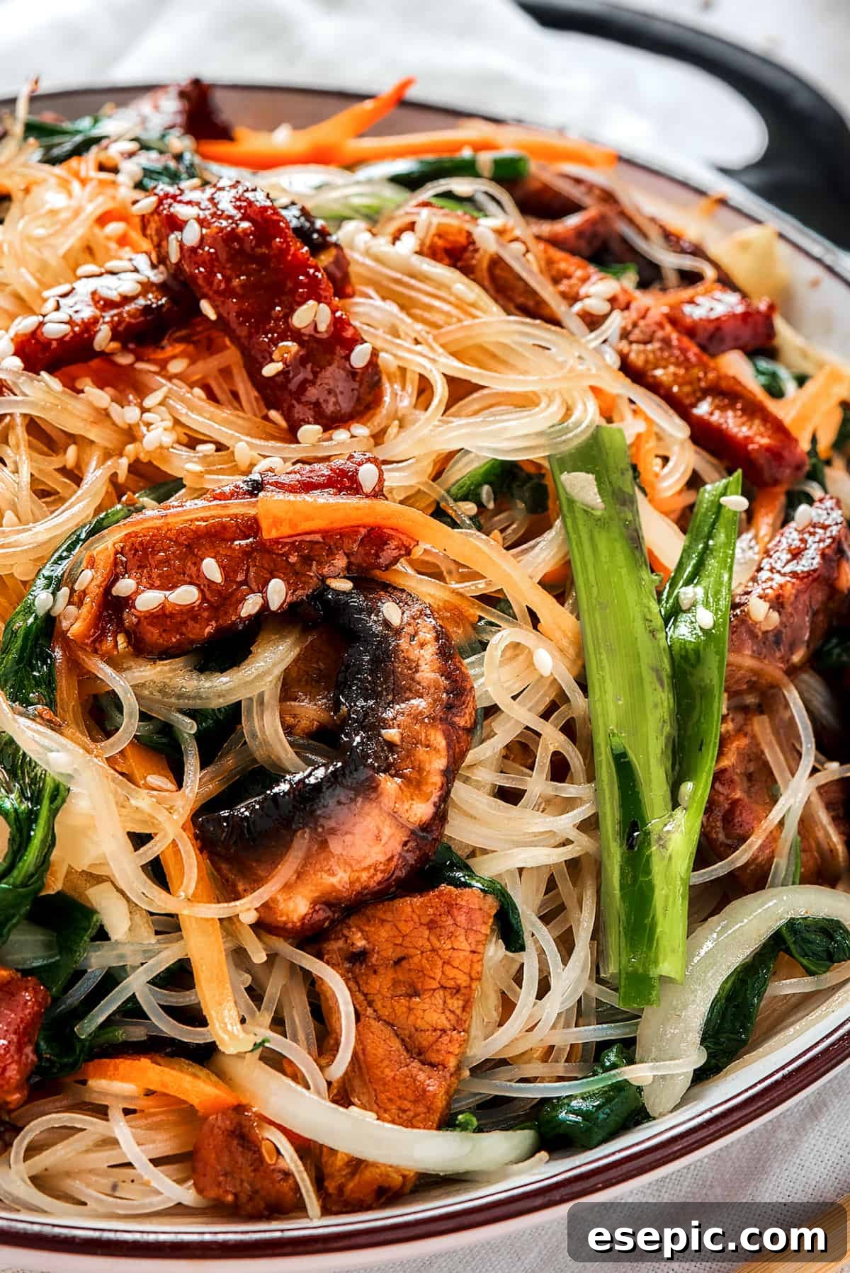 A shallow dish of beef noodle stir fry (Japchae), generously garnished with toasted sesame seeds, ready for serving.