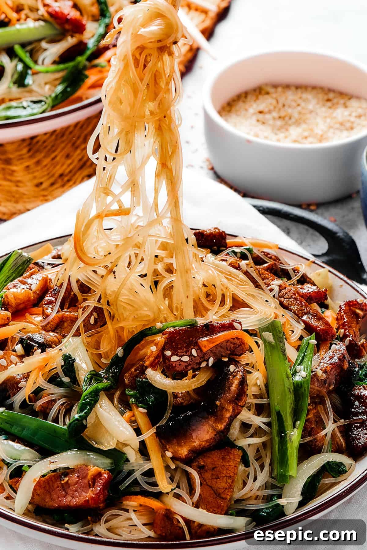 A bowl of beef stir fry with glass noodles (Japchae), with chopsticks pulling up a portion of the noodles, ready to be eaten.