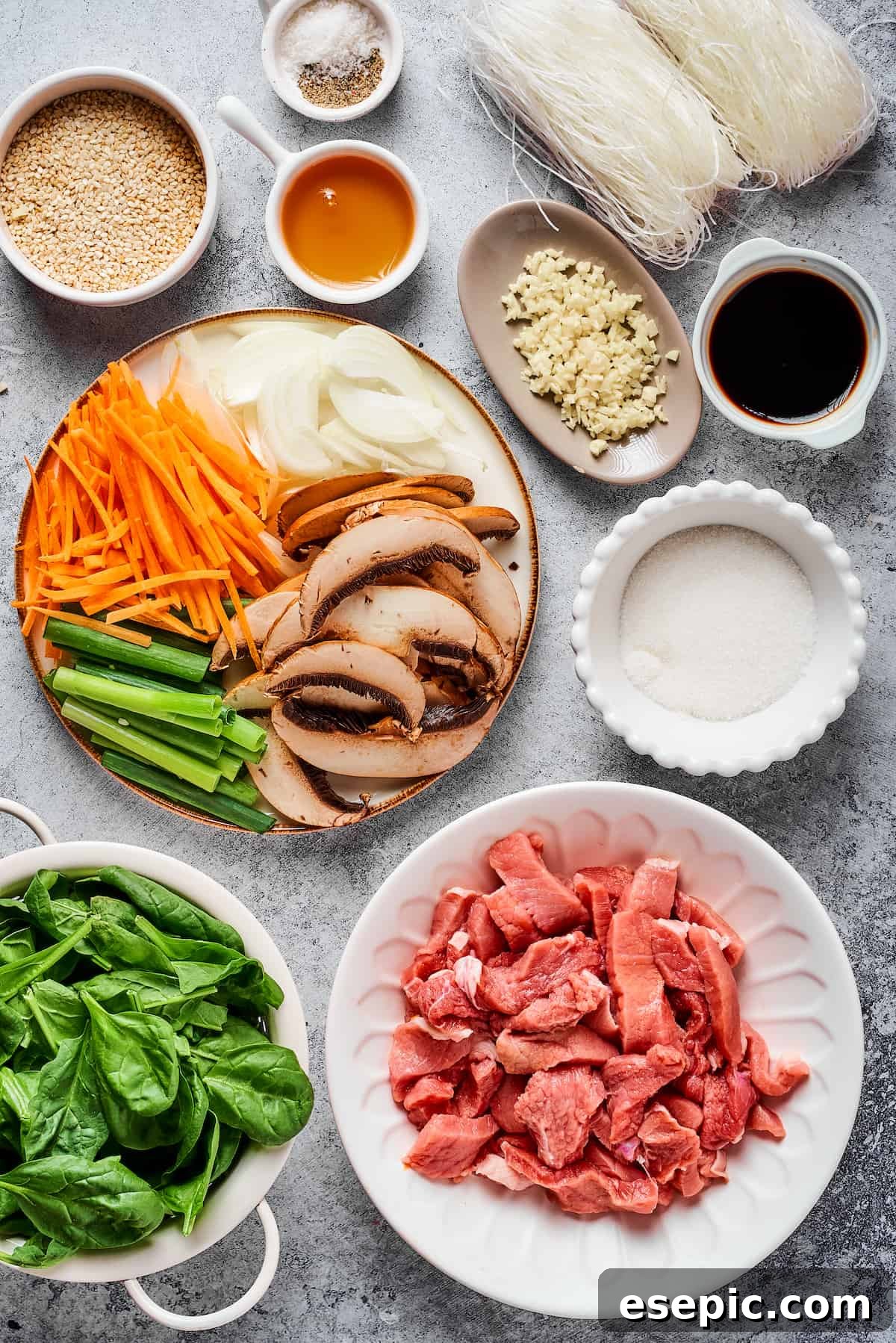 A vibrant flat lay of all the fresh ingredients for Beef Stir Fry with Glass Noodles (Japchae), including sliced steak, colorful vegetables like carrots, onions, green onions, mushrooms, and spinach, alongside essential seasoning components such as toasted sesame seeds, salt, pepper, dried glass noodles, sesame oil, garlic, and soy sauce.