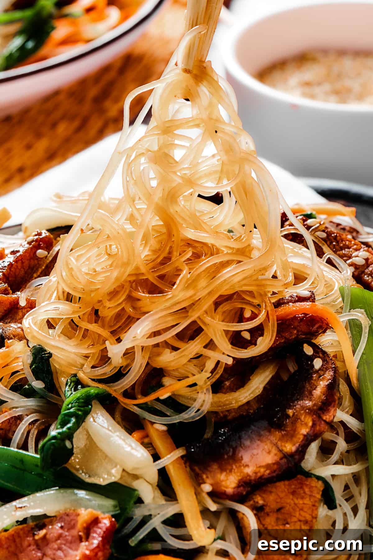 A perfectly plated bowl of beef stir fry with glass noodles (Japchae), with chopsticks lifting a tangle of noodles from the dish, highlighting their translucent and springy texture.