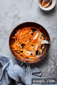 Matchstick carrots and thinly sliced onions being stir-fried in a skillet, showcasing the early stages of vegetable preparation for Japchae.