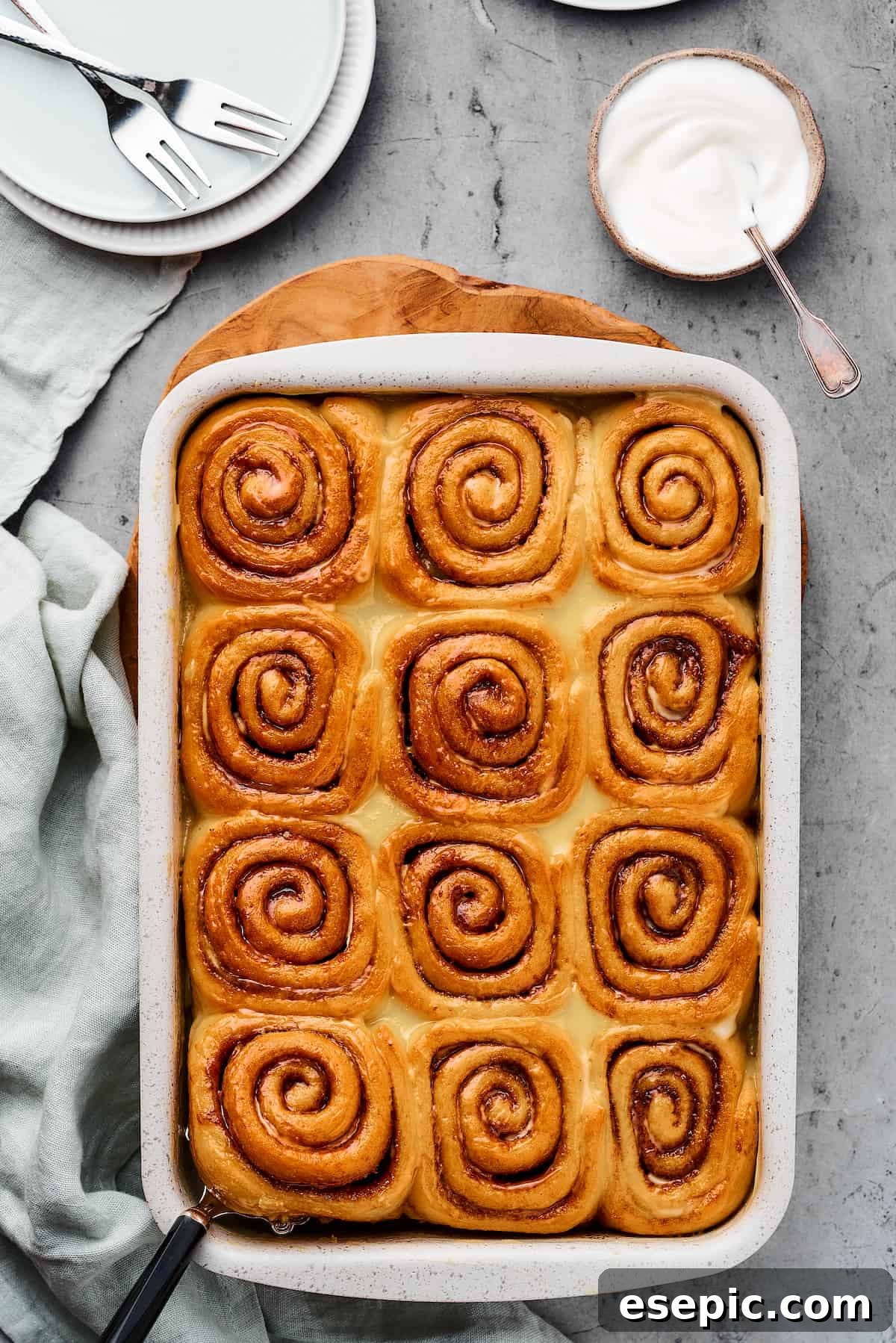 Freshly baked cinnamon rolls, golden brown on top, cooling in a baking pan, with a soft cloth dishtowel nearby.