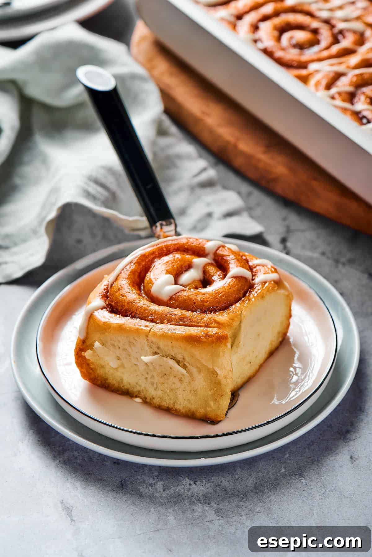 A single sticky cinnamon roll on a small white plate, accompanied by a fork and a neatly folded cloth napkin, inviting a delicious taste.