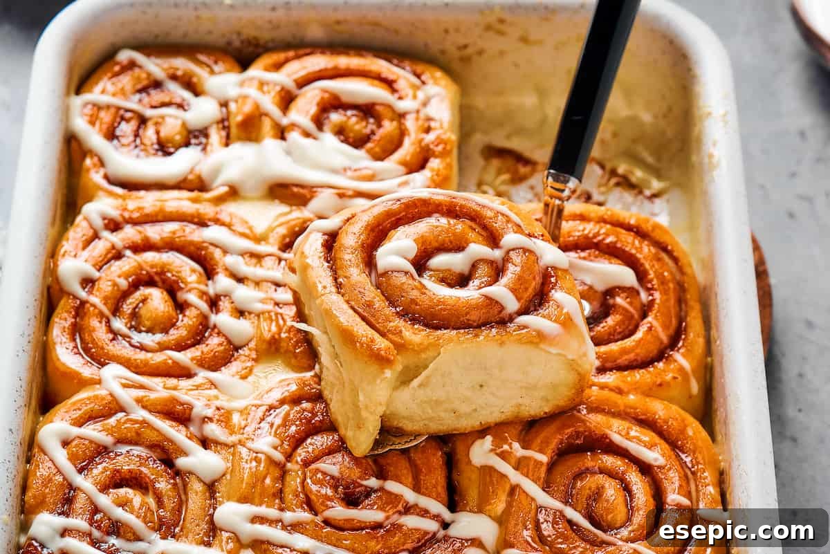 One sticky cinnamon roll, perfectly iced with a drizzle of glaze, resting gently on the pan with other baked rolls, showcasing its tempting texture.