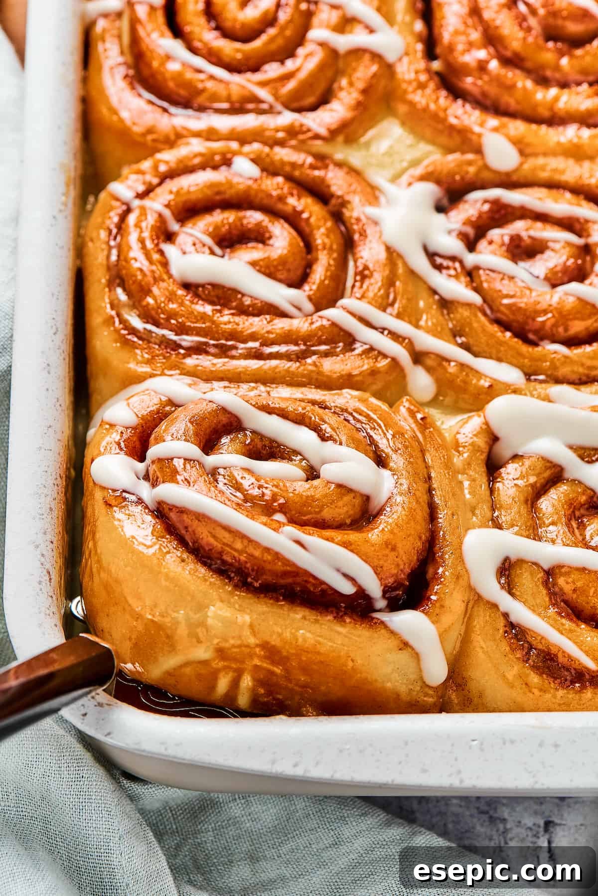 A pan of sticky homemade cinnamon buns, with one being carefully lifted from the corner using a metal spatula, showcasing its soft texture.