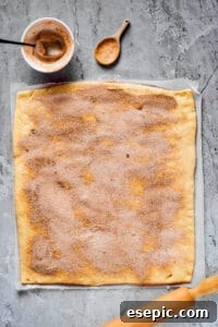 A work surface with a large rectangle of dough, a rolling pin, and a small bowl of cinnamon sugar nearby. Cinnamon sugar has been sprinkled generously all over the dough, ready for rolling.
