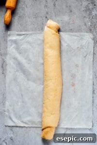 A long, tightly rolled-up piece of yeast dough, generously filled with cinnamon sugar, resting on a sheet of parchment paper, next to a rolling pin.