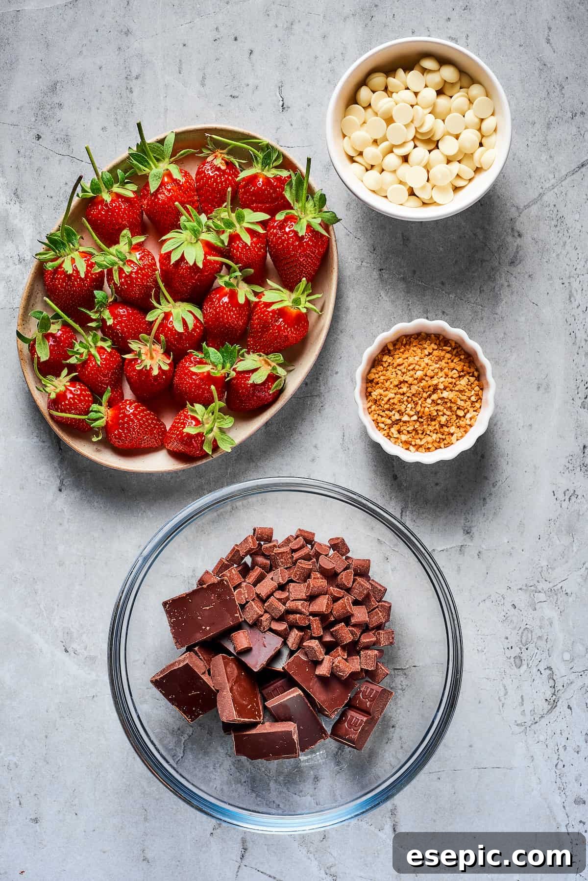 Effortless Chocolate Strawberry Delight 3 A flat lay photograph featuring fresh, vibrant strawberries with their green stems, a bowl of pristine white chocolate chips, a separate bowl of toasted, finely chopped hazelnuts, and a mix of chopped semisweet and milk chocolate chips ready for melting.