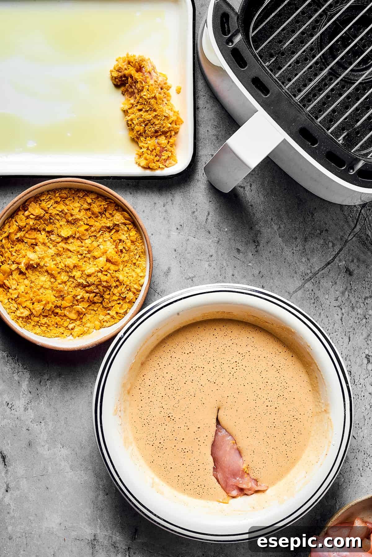 Utterly addictive, this easy recipe for Air Fryer Chicken Tempura is a favorite! Tender, juicy chicken strips are coated in a light and crispy batter, and then air-fried to golden perfection. A piece of chicken dipped in batter, next to a bowl of cornflakes. A breaded chicken strip is on an oiled baking sheet nearby, along with an open air fryer.