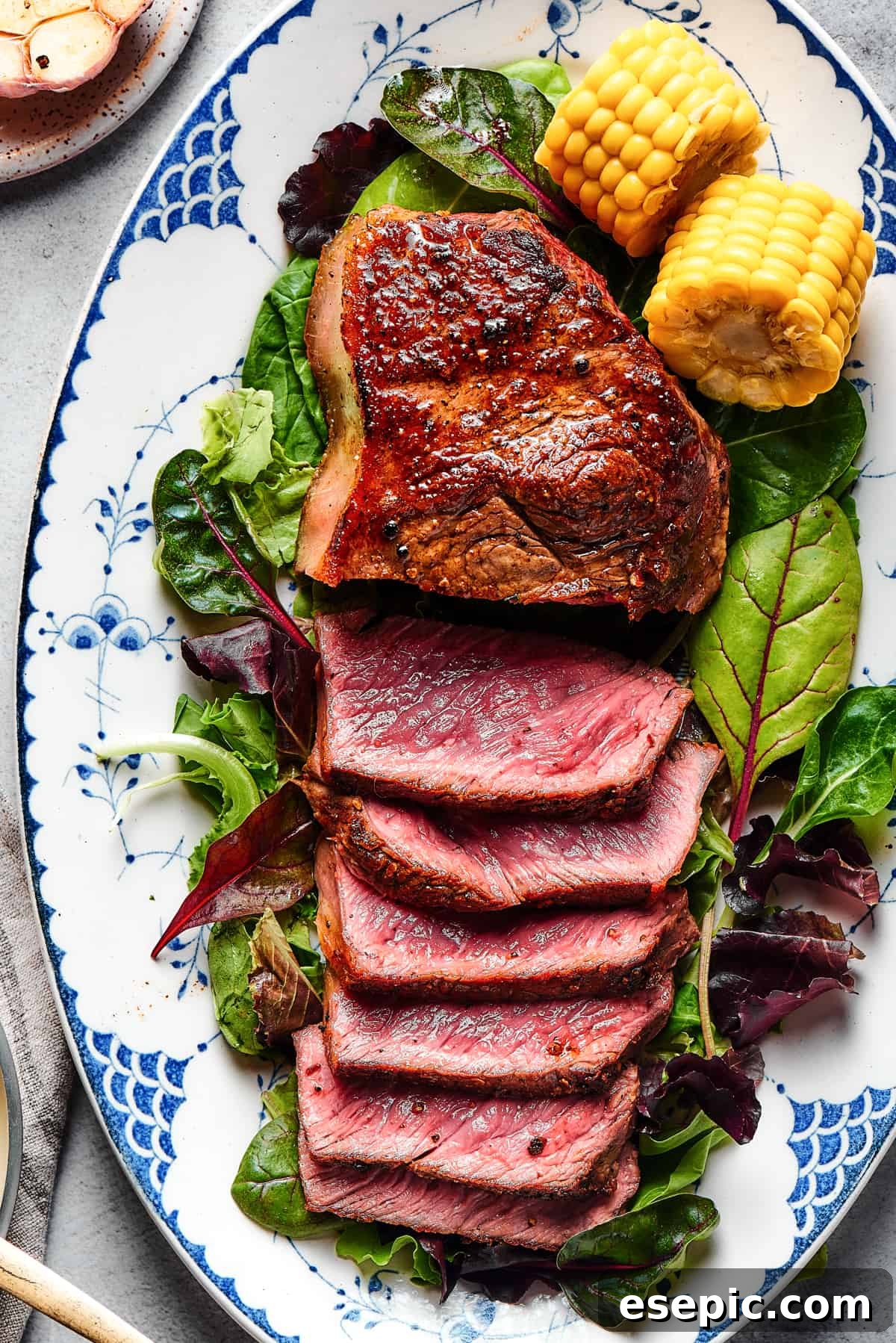 A stunning blue steak displayed on a bed of fresh greens, accompanied by corn on the cob. Half of the steak is expertly sliced and fanned out, magnificently showcasing its vibrant, ultra-rare interior.