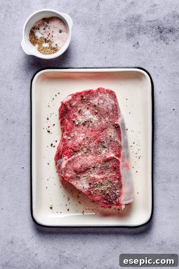 A thick cut of steak resting in an enamel pan, generously seasoned with coarse salt and freshly cracked black pepper.
