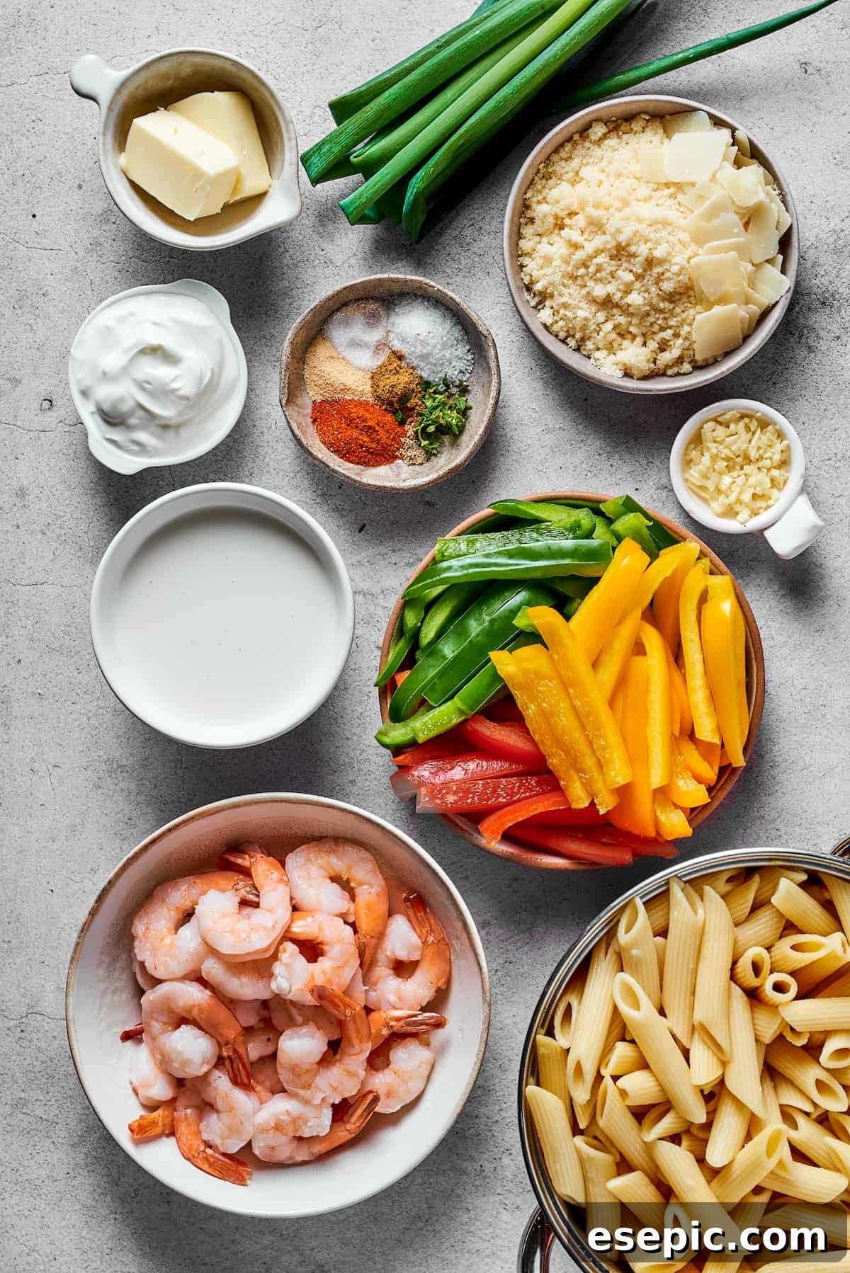 An array of Rasta Pasta ingredients laid out: Butter, green onions, parmesan cheese, cream cheese, various spices, garlic, heavy cream, sliced bell peppers (red, yellow, and green), shrimp, and penne pasta.