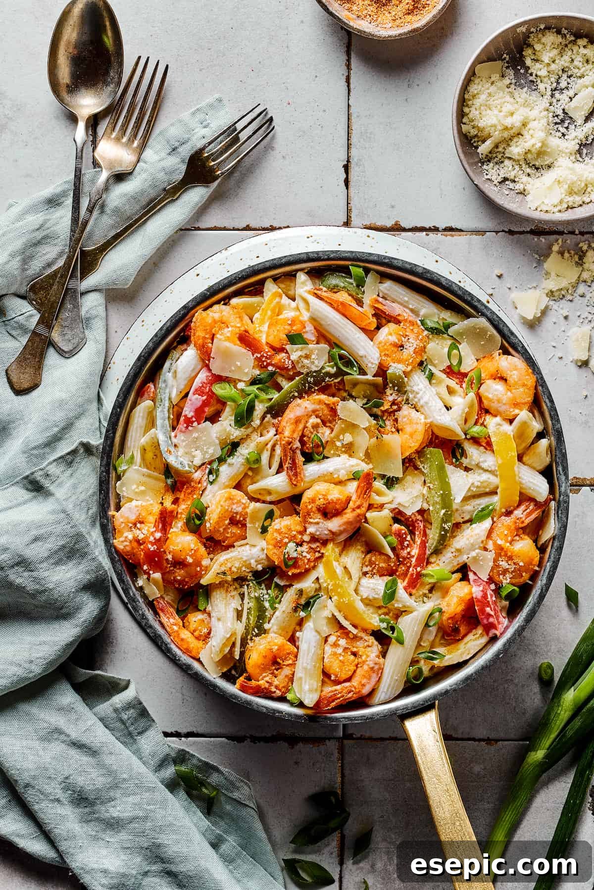 A beautifully presented skillet of Rasta Pasta with shrimp, penne, and colorful vegetables, garnished with green onions, ready to be enjoyed. Forks and spoons are placed nearby on a cloth napkin.