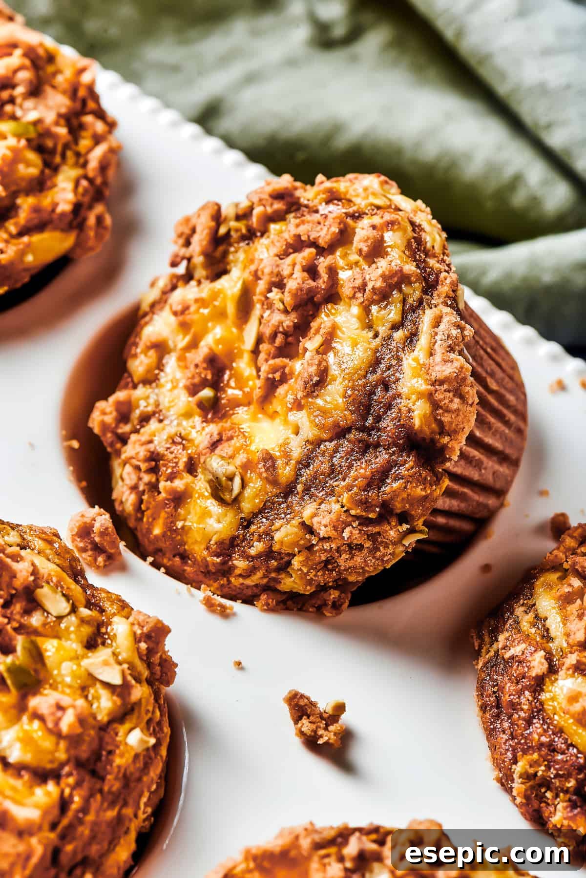 Pumpkin Cream Cheese Muffins: Spiced pumpkin cake with a smooth cream cheese swirl and buttery streusel topping. A muffin tin with one pumpkin cream cheese muffin turned diagonally in its well, with small pieces of streusel topping scattered around it.