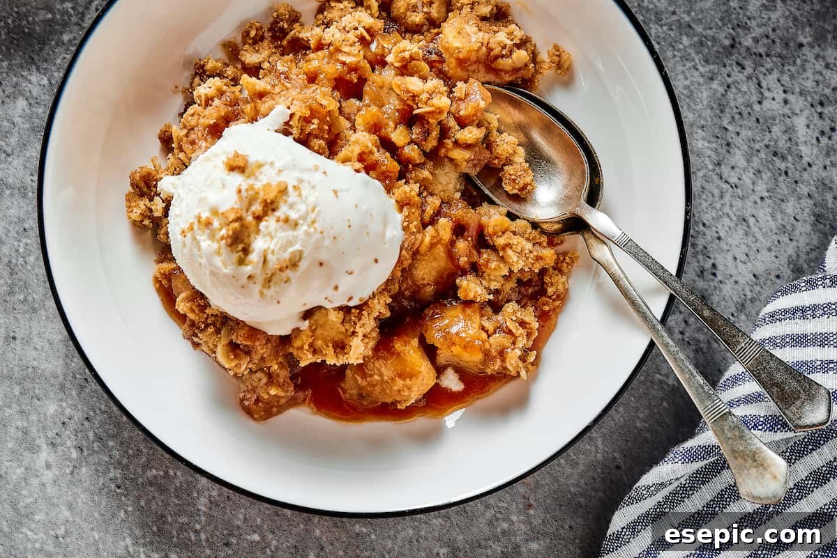 A serving of homemade apple crisp in a dessert bowl, generously topped with a scoop of vanilla ice cream and drizzled with caramel, set on a dark surface.
