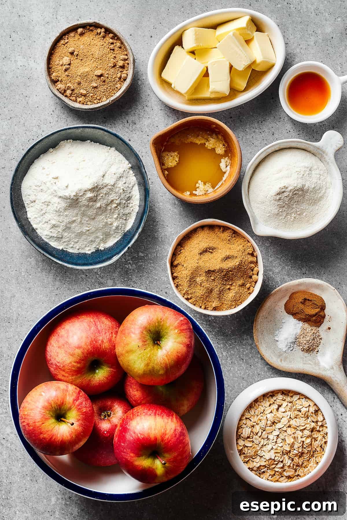 An organized display of fresh ingredients for apple crisp: brown sugar, butter, vanilla extract, flour, melted butter, spices, sliced apples, and rolled oats.