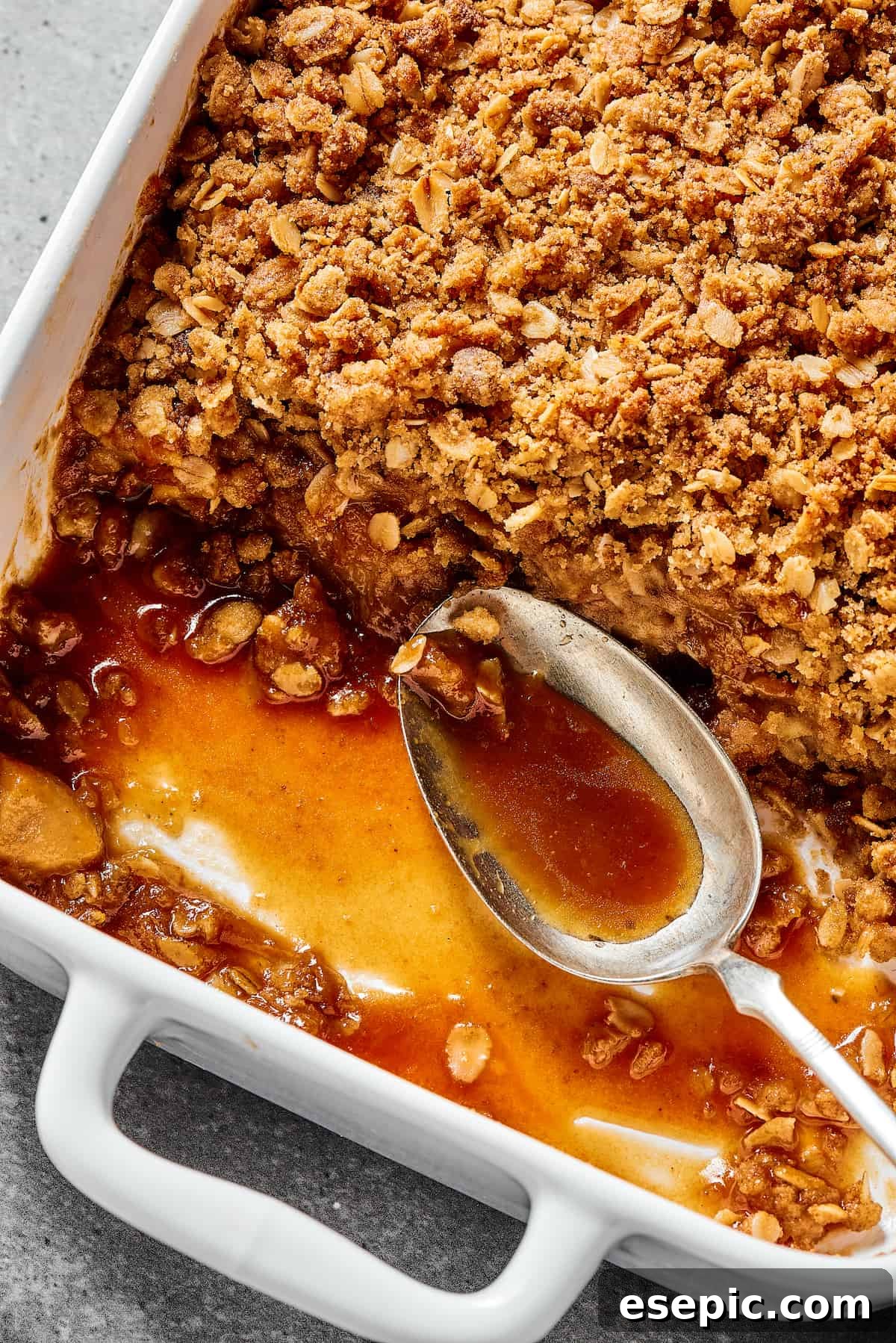 A close-up view of a freshly baked apple crisp in a white casserole dish, showing the golden oat topping and bubbling apple filling around the edges, with a serving spoon resting nearby.
