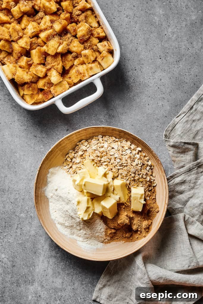 Next to a baking dish filled with seasoned apples, a bowl holds the ingredients for the crisp topping: rolled oats, cubed cold butter, and flour, ready to be combined.