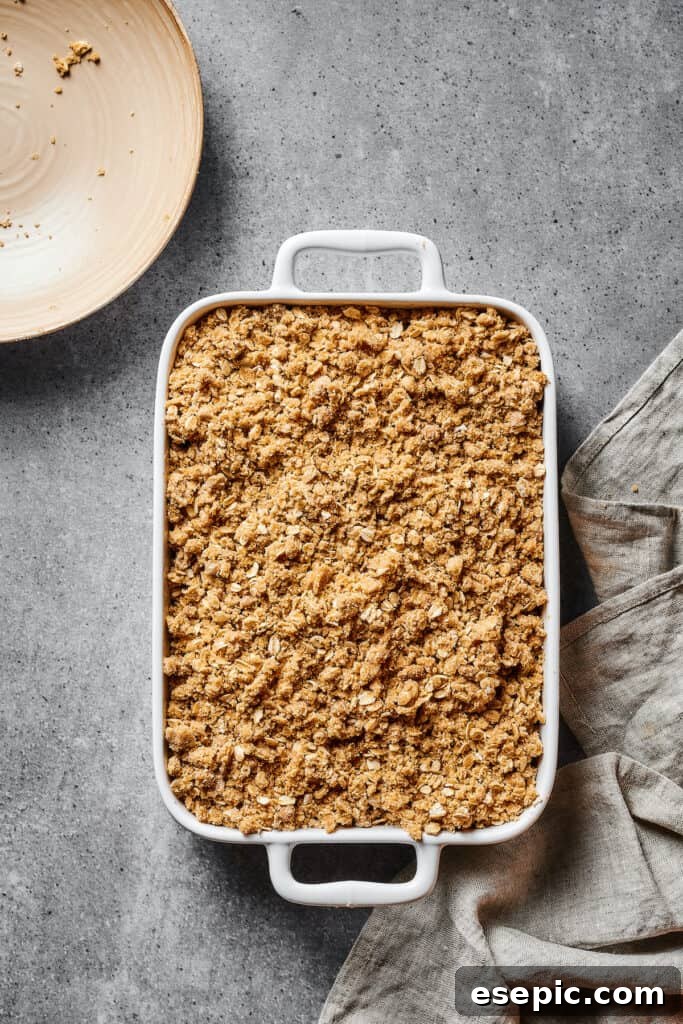 An unbaked apple crisp sits in a white casserole dish, showcasing the generous, uniform layer of crumbly oat topping over the apple filling, ready for the oven.