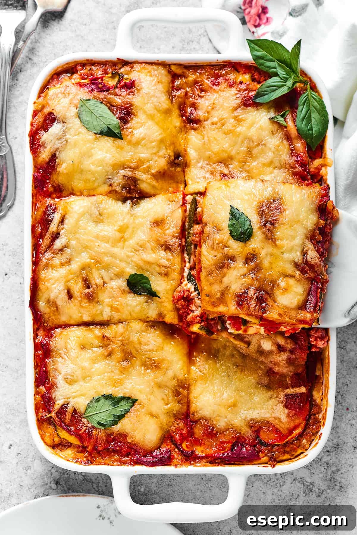 Comforting Zucchini Lasagna with meat sauce is the perfect low-carb family meal. Cheesy, saucy, and healthy, with zucchini instead of pasta! A slice of cheesy zucchini lasagna being lifted from a white rectangular baking dish, revealing its delicious layers.