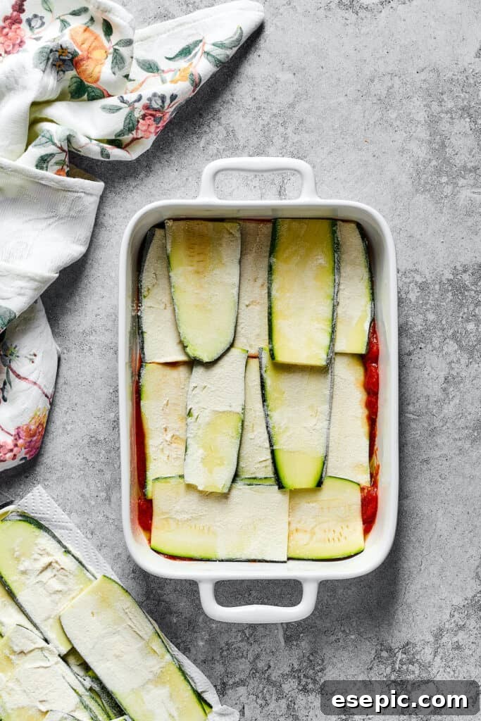 Floured zucchini strips carefully layered over the tomato sauce base in a baking dish, forming the first 'noodle' layer.