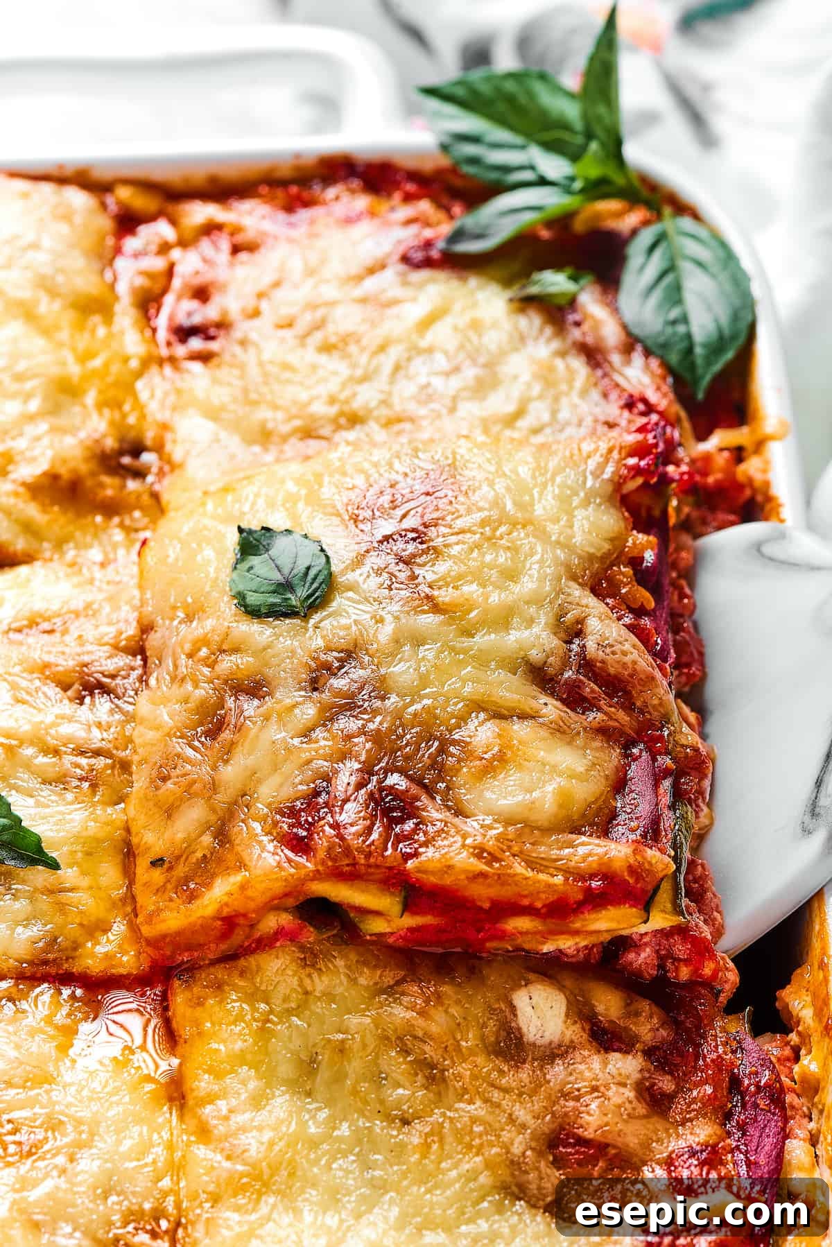 Comforting Zucchini Lasagna with meat sauce is the perfect low-carb family meal. Cheesy, saucy, and healthy, with zucchini instead of pasta! A large portion of Zucchini Lasagna being carefully lifted out of a white rectangular baking dish, ready to be served.