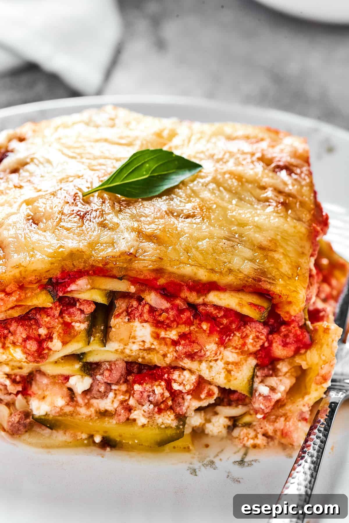 Comforting Zucchini Lasagna with meat sauce is the perfect low-carb family meal. Cheesy, saucy, and healthy, with zucchini instead of pasta! A generous serving of Zucchini Lasagna neatly placed on a white dinner plate, accompanied by a fork, ready to be enjoyed.