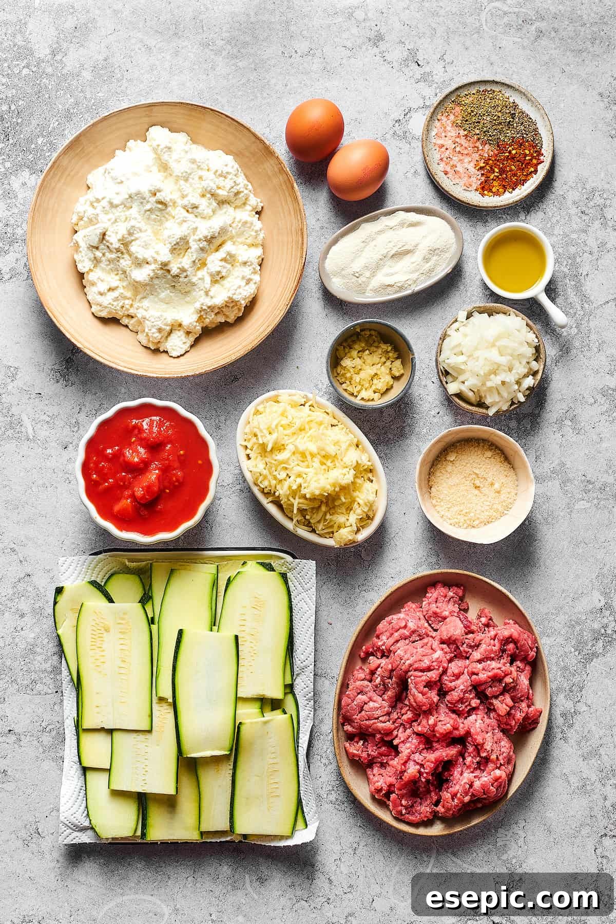 Comforting Zucchini Lasagna with meat sauce is the perfect low-carb family meal. Cheesy, saucy, and healthy, with zucchini instead of pasta! Individual ingredients for Zucchini Lasagna arranged neatly. From top left: Cottage cheese, eggs, various seasonings, flour, olive oil, minced garlic, diced onion, crushed tomatoes, shredded mozzarella, grated Parmesan, zucchini slices, and ground beef.