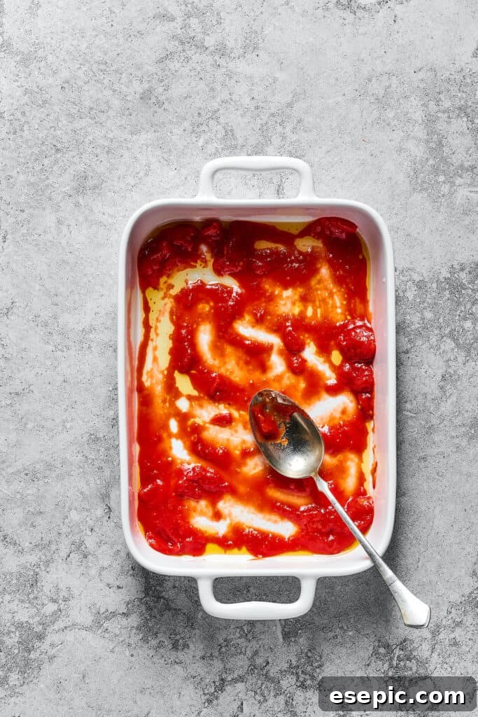 A generous layer of tomato sauce spread evenly across the bottom of a white rectangular baking dish, preparing the base for lasagna.