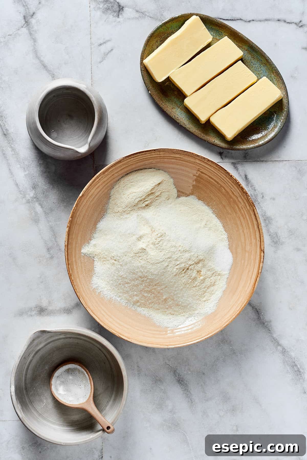 A mixing bowl with dry baking ingredients in it. Arranged around the mixing bowl are dishes of butter and ice water.