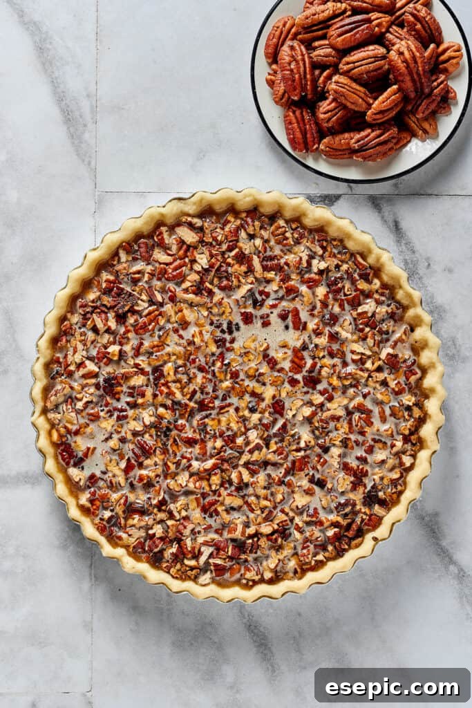 An overhead view of a partially assembled pecan pie in a crust, with chopped pecans lining the bottom and the maple filling poured over them, awaiting the decorative pecan halves.