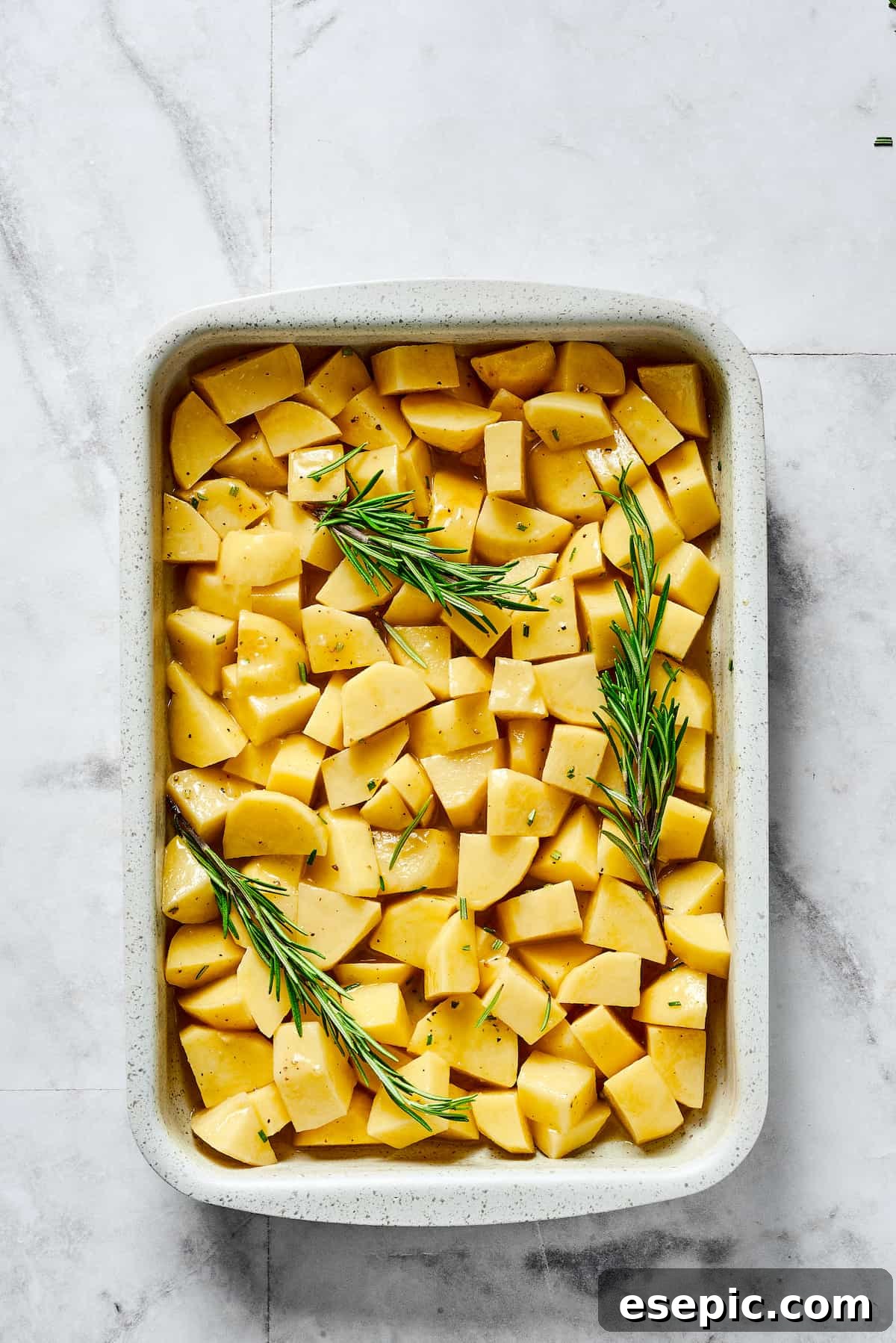 Seasoned raw potatoes and rosemary sprigs spread evenly in a baking dish before roasting.