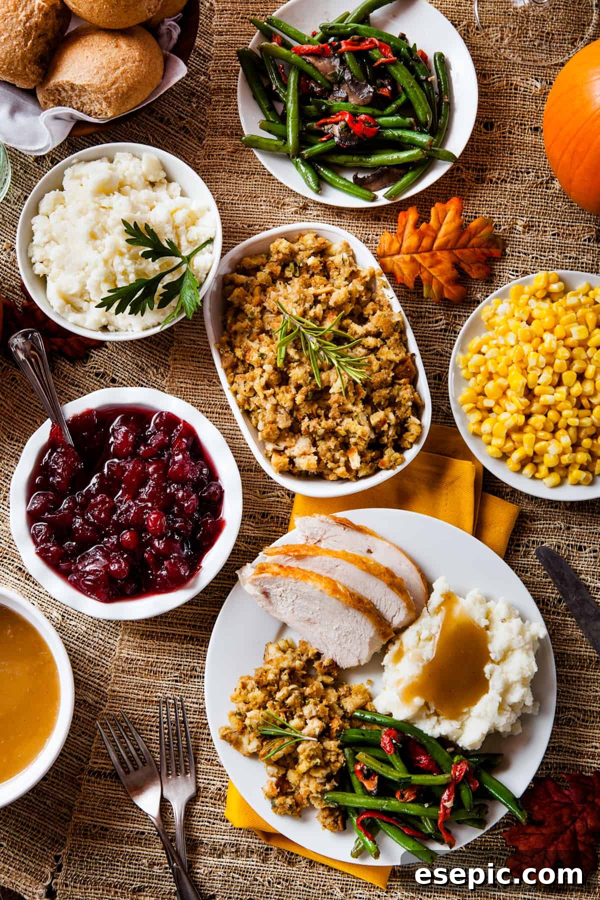 An overhead shot of a holiday spread, featuring a gravy boat, cranberry sauce, mashed potatoes, stuffing, green beans, corn, and a plate with sliced turkey and sides.