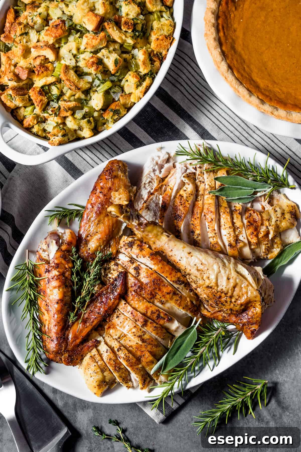 An overhead shot of a festive holiday table featuring a carved roast turkey, alongside bowls of stuffing and a slice of pumpkin pie.