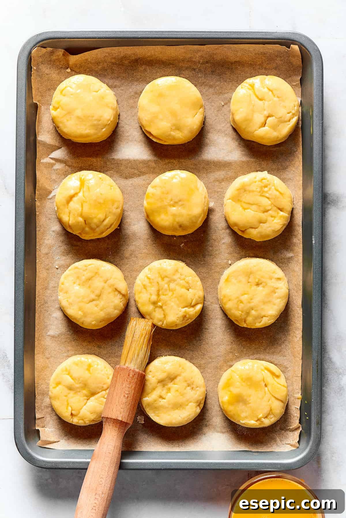 A dozen unbaked biscuits on a baking sheet, being brushed with beaten egg.