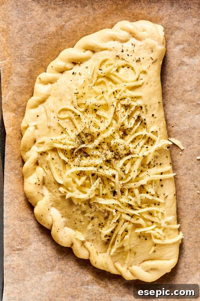 An unbaked calzone, brushed with egg wash and topped with shredded cheese and Italian seasoning.