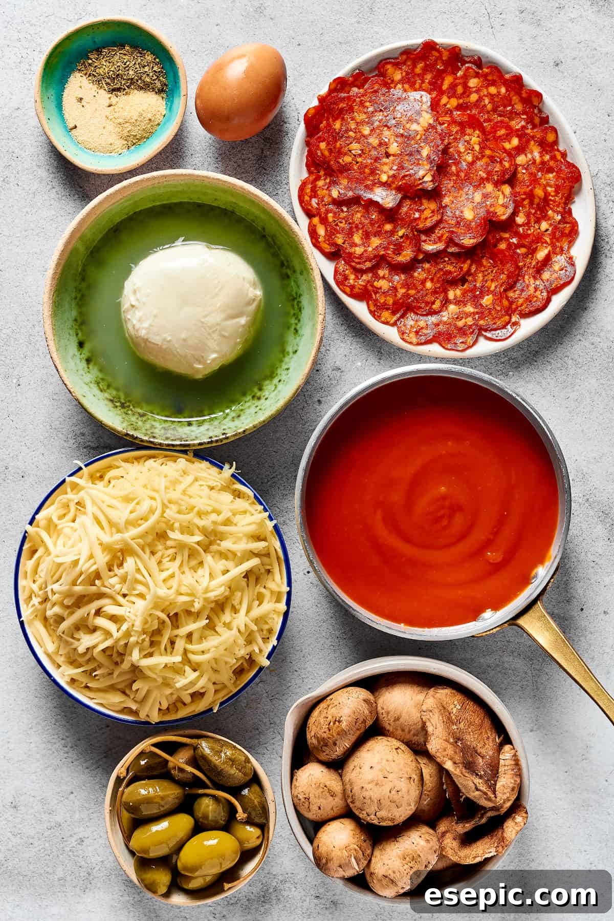 Various calzone filling ingredients laid out: Seasonings, an egg, pepperoni, fresh mozzarella, pizza sauce, shredded cheese, olives and capers, sliced mushrooms.