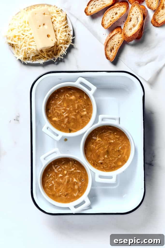 Individual oven-safe crocks filled with French onion soup, with toasted baguette slices and shredded Gruyere and Swiss cheese waiting to be assembled.