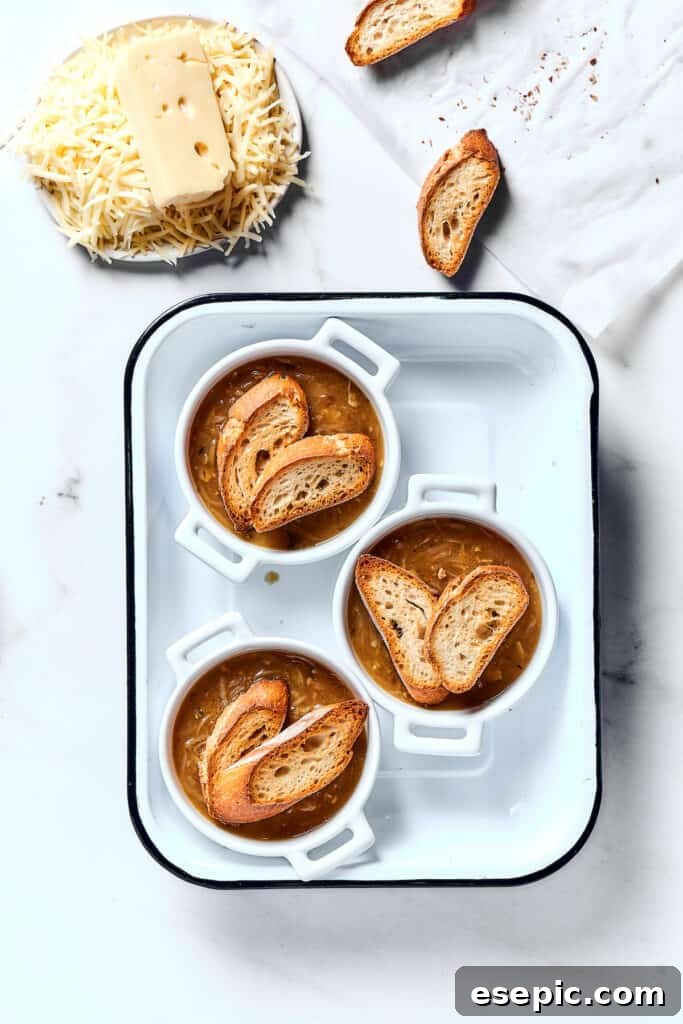Individual oven-safe bowls of French onion soup topped with toasted baguette slices, awaiting the cheese layer.