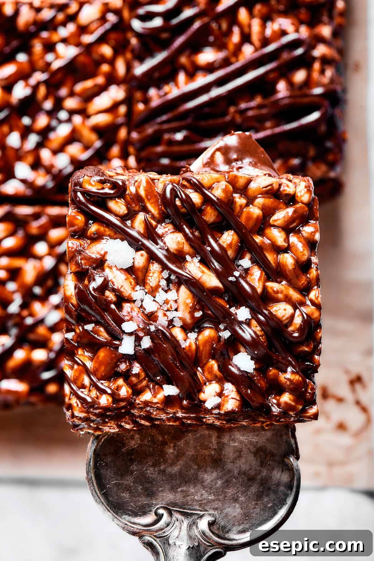 Perfectly crafted chocolate rice krispie treat, ready to be served. A chocolate cereal bar square being lifted from a platter with a silver dessert server, highlighting its gooey texture.