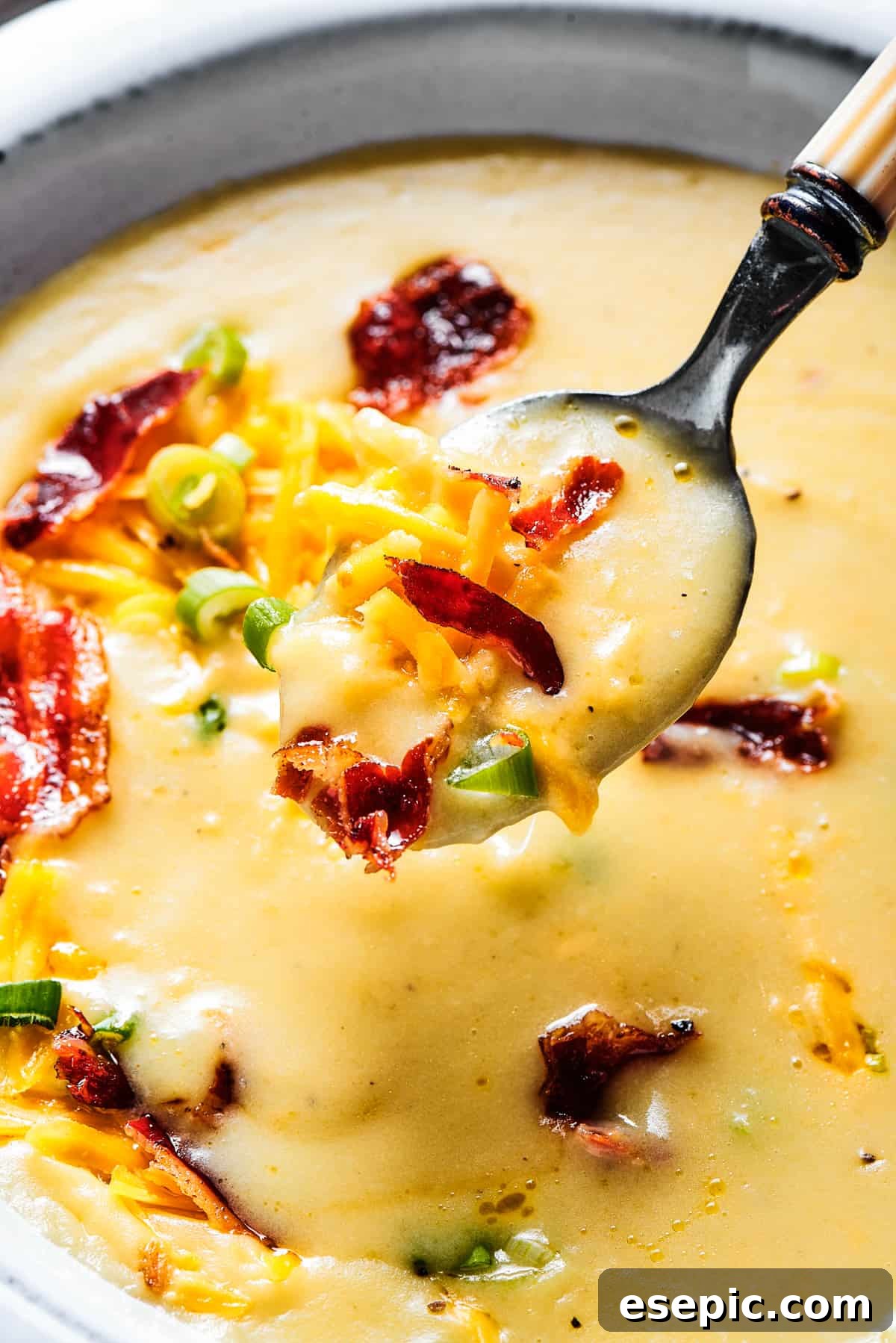 A beautifully presented white bowl of rich, creamy loaded potato soup, with a spoon gently lifted out, showcasing the thick texture and delicious toppings.