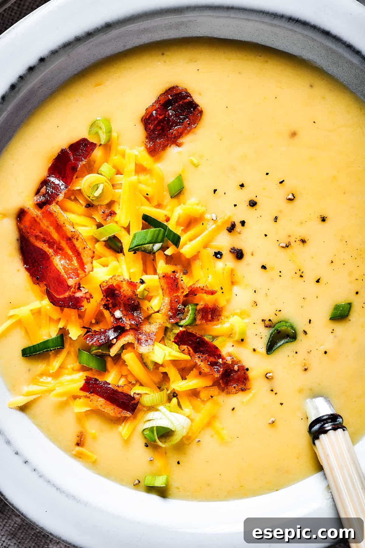 A close-up view of a bowl of creamy potato soup, garnished lavishly with crispy bacon bits, vibrant chopped green onions, and melting shredded cheese, highlighting its rich texture and enticing toppings.