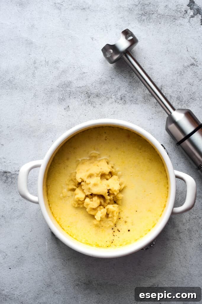 A large pot filled with creamy potato soup and mashed potatoes, with an immersion blender resting on the counter nearby, ready for blending.