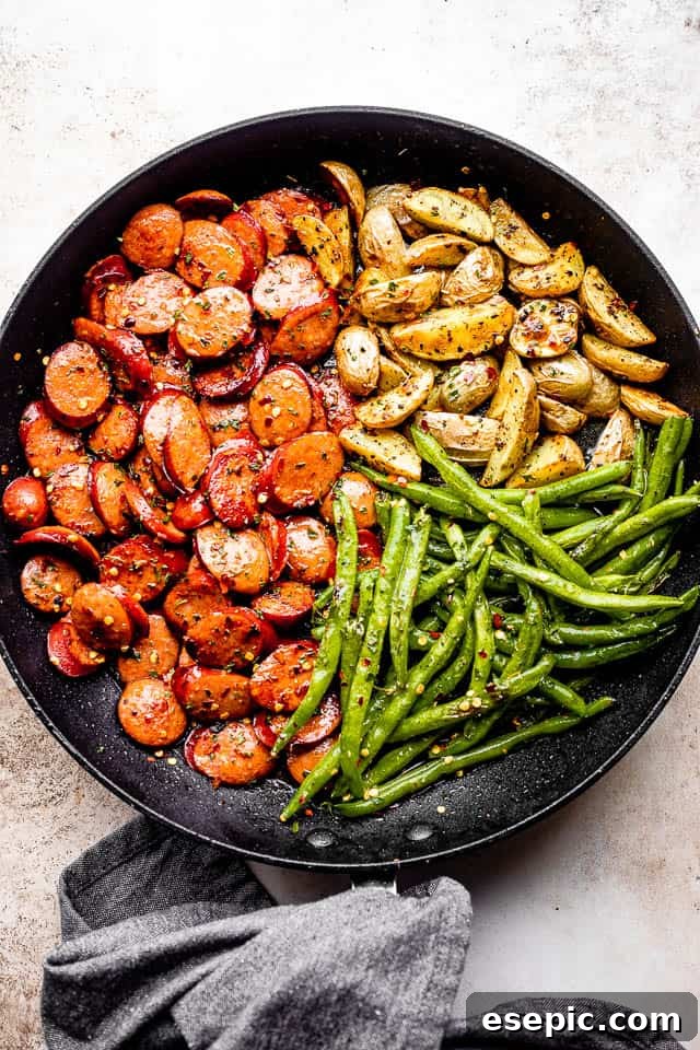 A rustic black skillet filled with slices of golden-brown smoked sausage, tender roasted potatoes, and vibrant green beans, resting on a grey linen kitchen towel, evoking a hearty and quick meal.