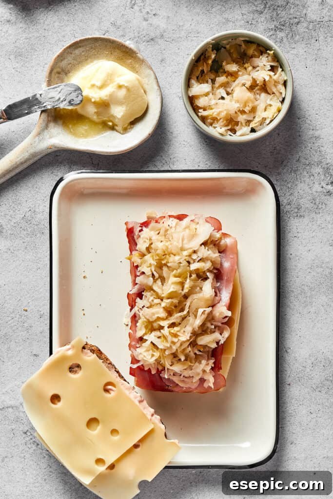 One slice of bread topped with cheese, corned beef, and sauerkraut, next to another slice with just cheese.