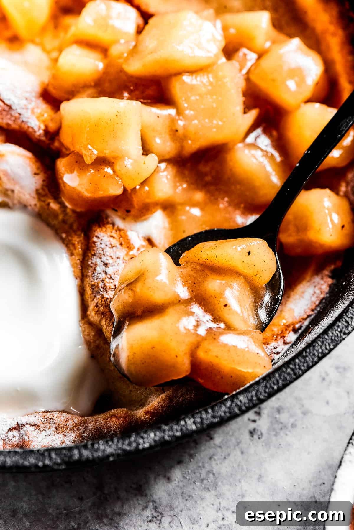 Spooning warm homemade apple pie filling on top of a fluffy Dutch baby pancake, ready for a delightful breakfast or dessert.