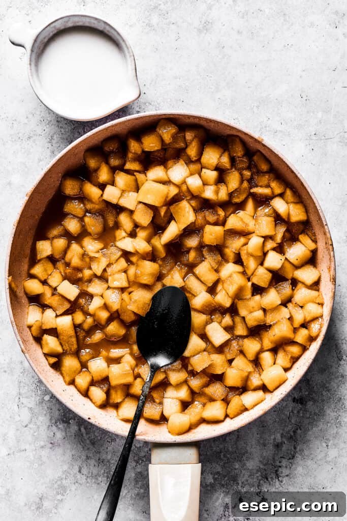 Diced apples cooking gently in a skillet, absorbing the rich flavors of butter, sugar, and spices, with a dark spoon resting inside.