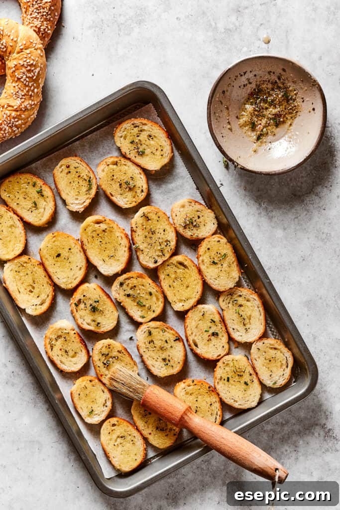 Bagel rounds neatly lined up on a baking sheet, brushed with seasoned butter. A pastry brush rests beside a dish of butter.