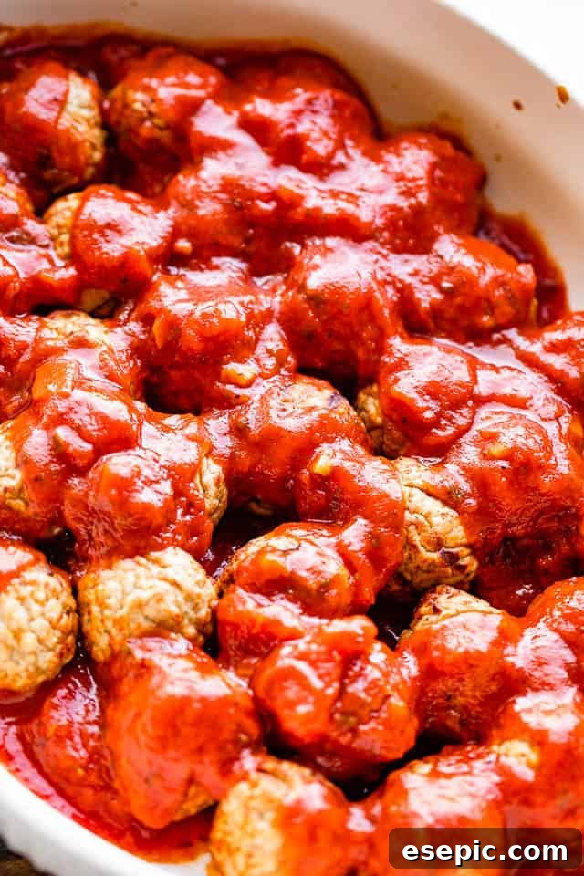 Cheesy baked meatball casserole with mini meatballs topped with your favorite marinara and loads of melty cheese! An easy dinner idea! A hand pouring rich marinara sauce over perfectly arranged, partially baked meatballs in a white casserole dish, preparing for the next baking stage.